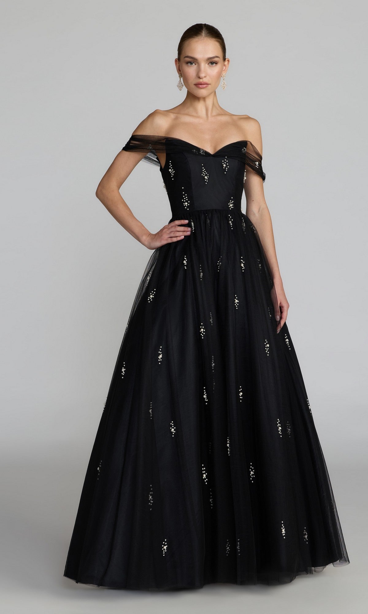 A woman wearing the Mac Duggal 13227 long formal dress in black, featuring an off-the-shoulder tulle ballgown design with small white embellishments, stands against a plain light gray background.